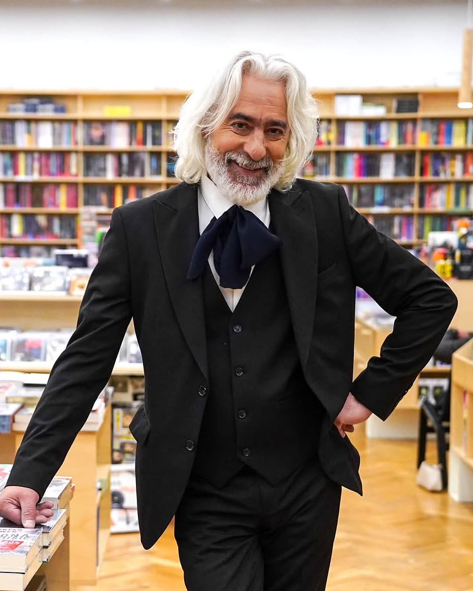 Dumitru Chicu, zâmbind, în costum negru, sprijinit de o masă cu cărți într-o librărie